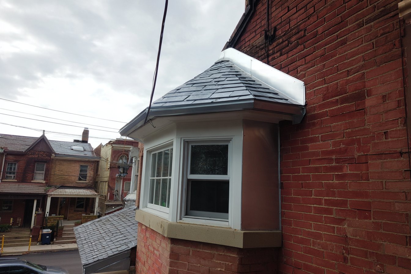 Small shingled roof over a bay window on a Queens brick row house, with new metal flashing tucked into the wall where the bay roof meets the house