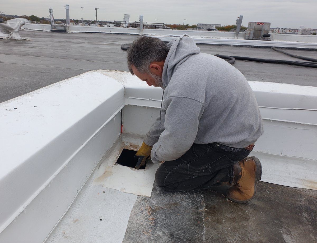 Hearth Roofing roofer kneeling at an interior roof drain on a Queens commercial flat roof, checking the membrane seam around it